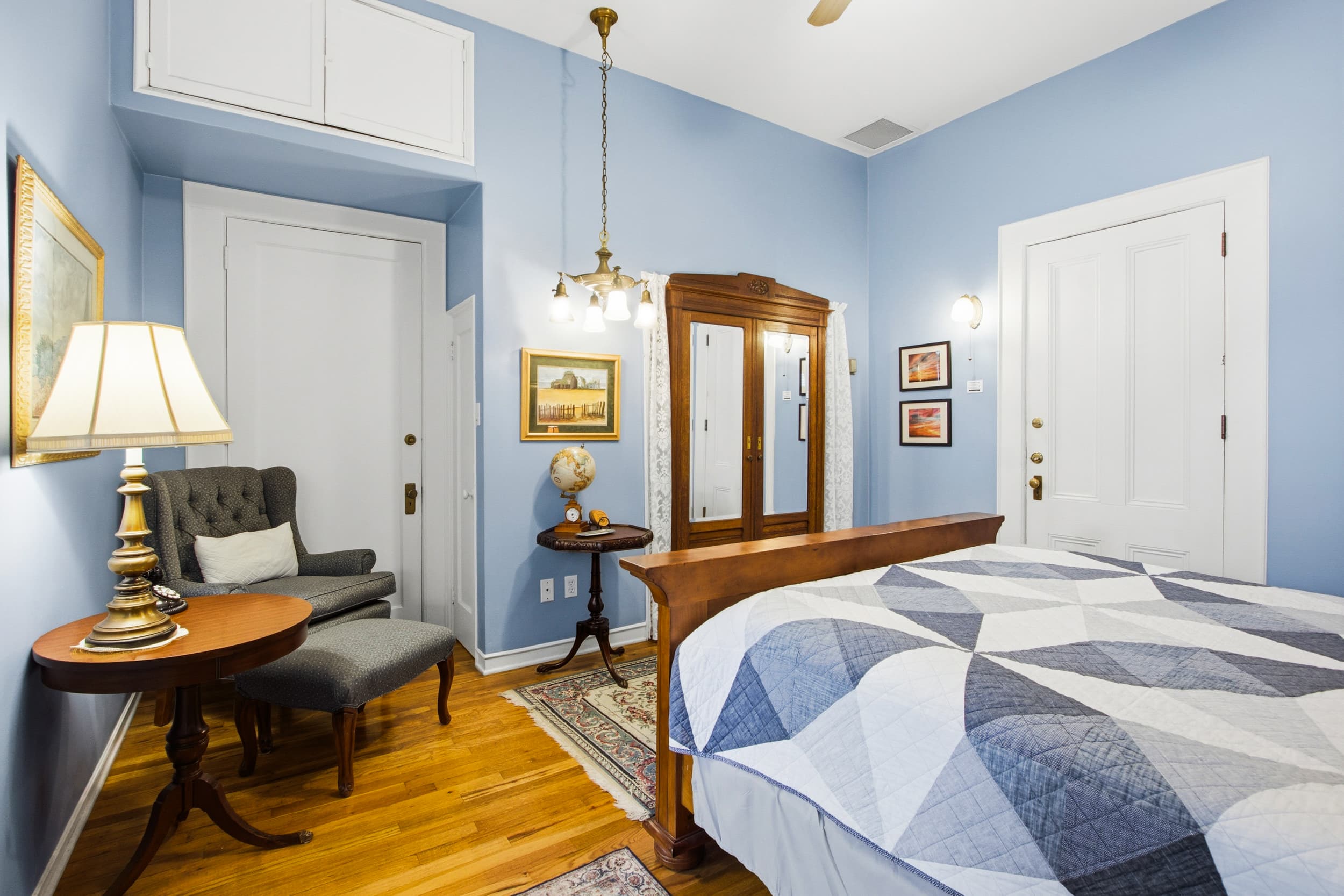 A cozy bedroom featuring a blue wall, a wooden bed with a patterned quilt, a lamp, a chair, and a vintage mirror.