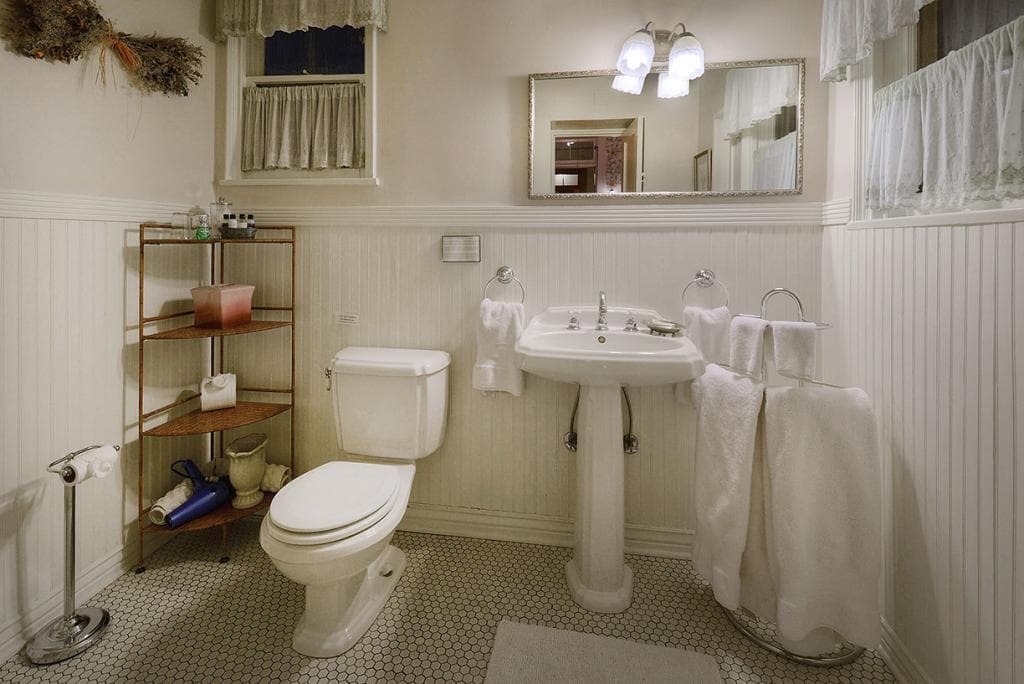 Brightly lit, vintage-style bathroom featuring a sink, toilet, towels, and a wooden shelf with toiletries.
