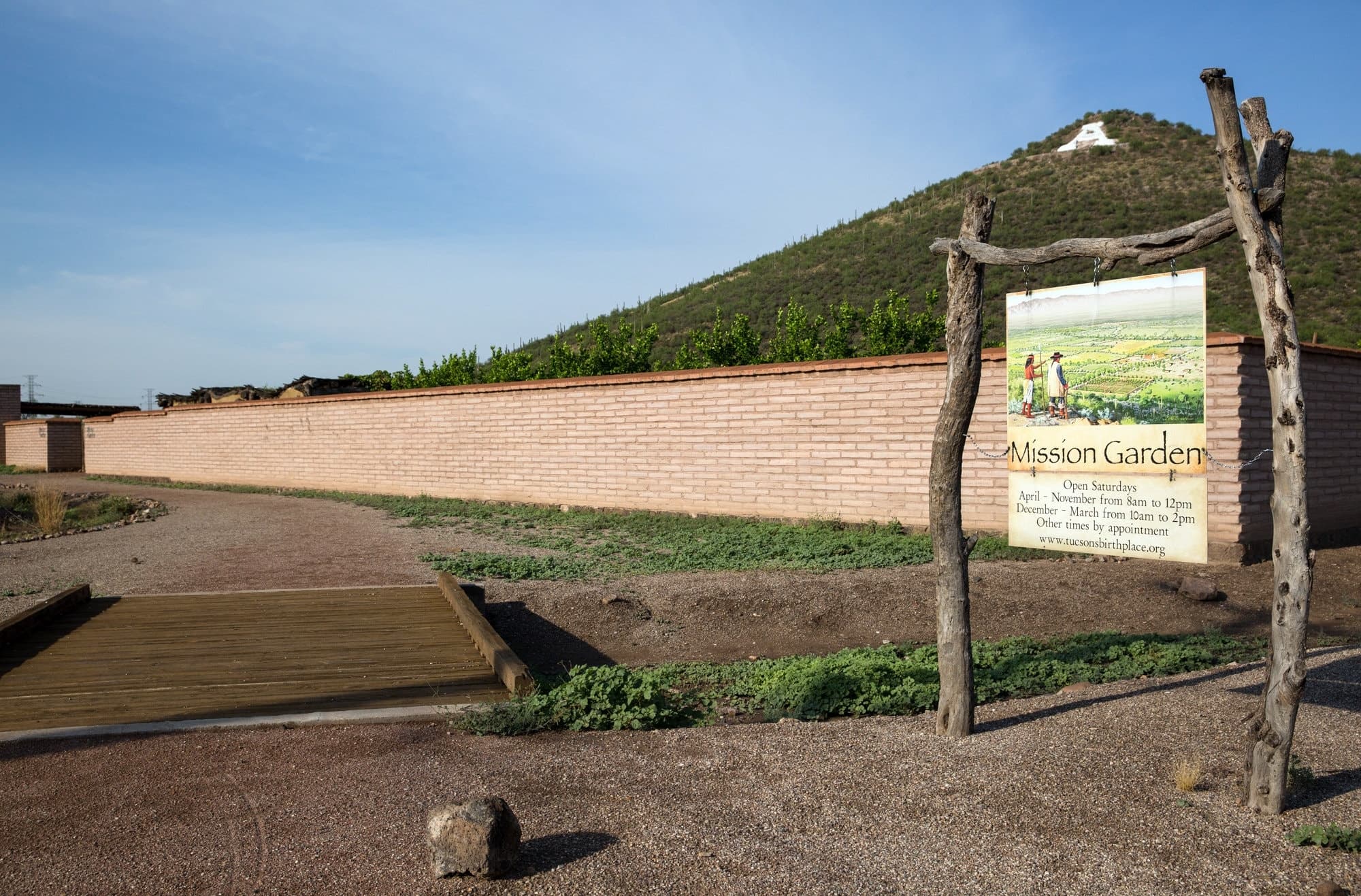 A wooden sign for "Mission Garden" stands near a path surrounded by greenery and a hillside in the background.