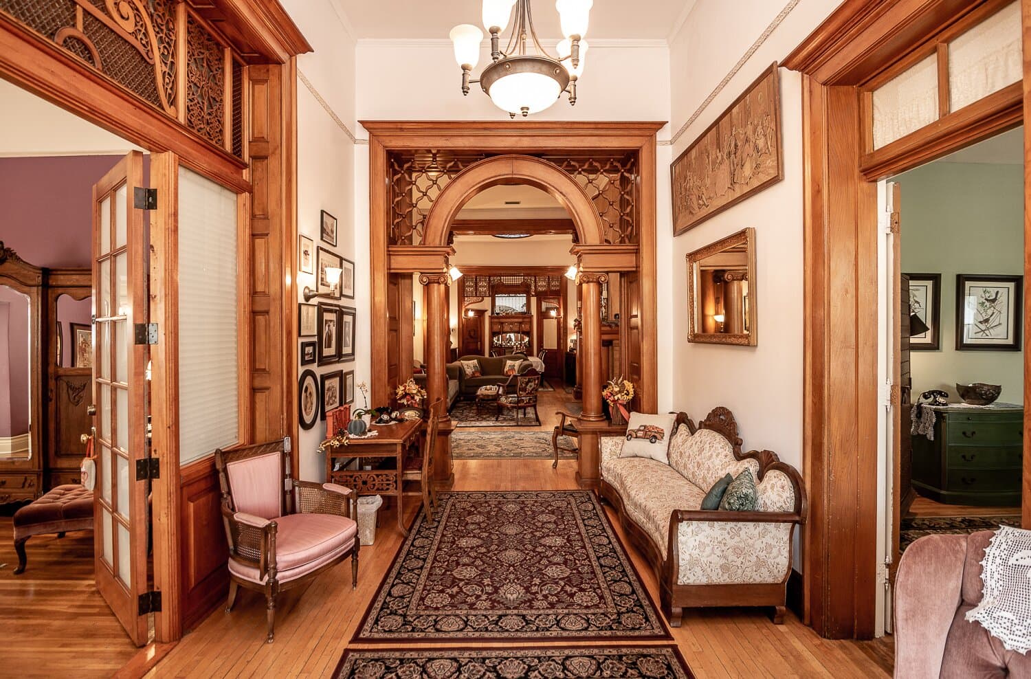 A spacious hallway with ornate woodwork, a patterned rug, and vintage seating leading to a cozy living area.