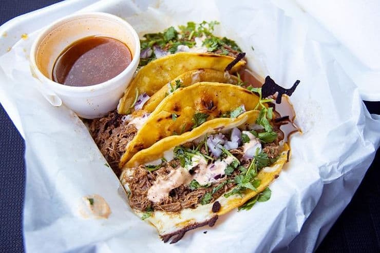 Three tacos filled with shredded meat, topped with onions and cilantro, served with a side of sauce.