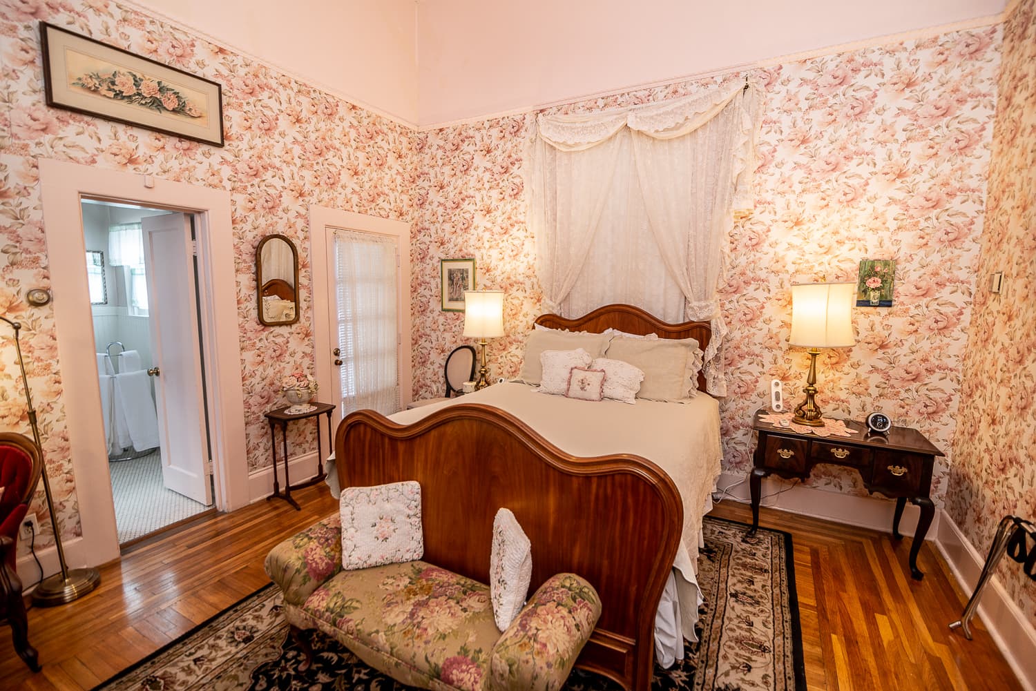 A cozy bedroom with floral wallpaper, an antique bed, and a small sitting area.