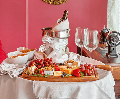 A wooden cheese platter with fruits and nuts, accompanied by a wine bottle in an ice bucket and two glasses, set against a pink wall.