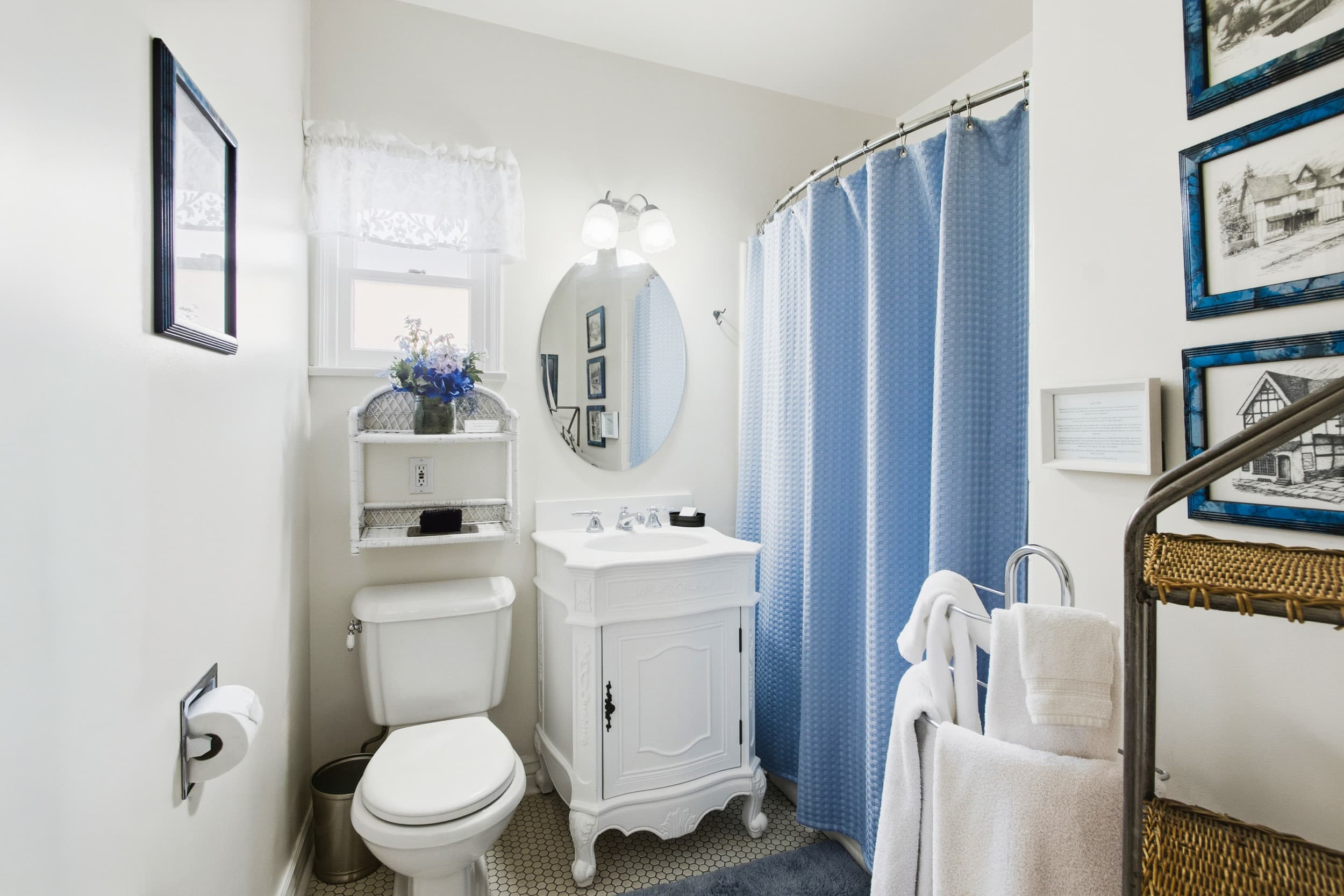A small, bright bathroom featuring a white vanity, a round mirror, a blue shower curtain, and decorative wall art.