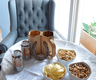 A cozy table setting featuring two cans of beer, a wooden mug holder, and bowls of snacks including chips, nuts, and crackers, with a plush armchair in the background.