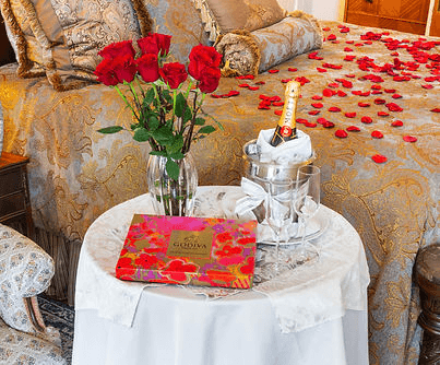 A romantic setting featuring a vase of red roses, a bottle of champagne in an ice bucket, two glasses, and a box of chocolates on a small table beside a luxurious bed strewn with rose petals.
