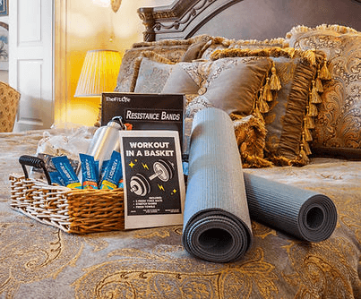 A cozy bed with decorative pillows, featuring a basket containing resistance bands, a water bottle, and workout instructions, alongside two exercise mats.