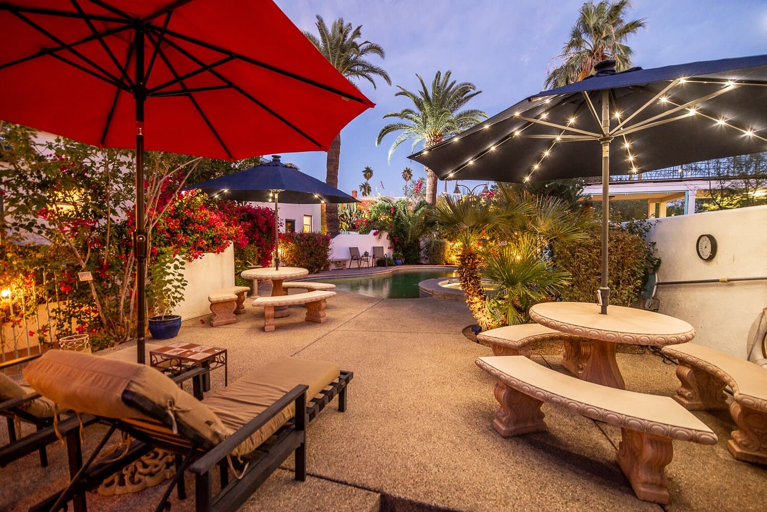 A cozy outdoor space featuring lounge chairs, tables, and umbrellas, surrounded by lush greenery and a pool under evening lights.