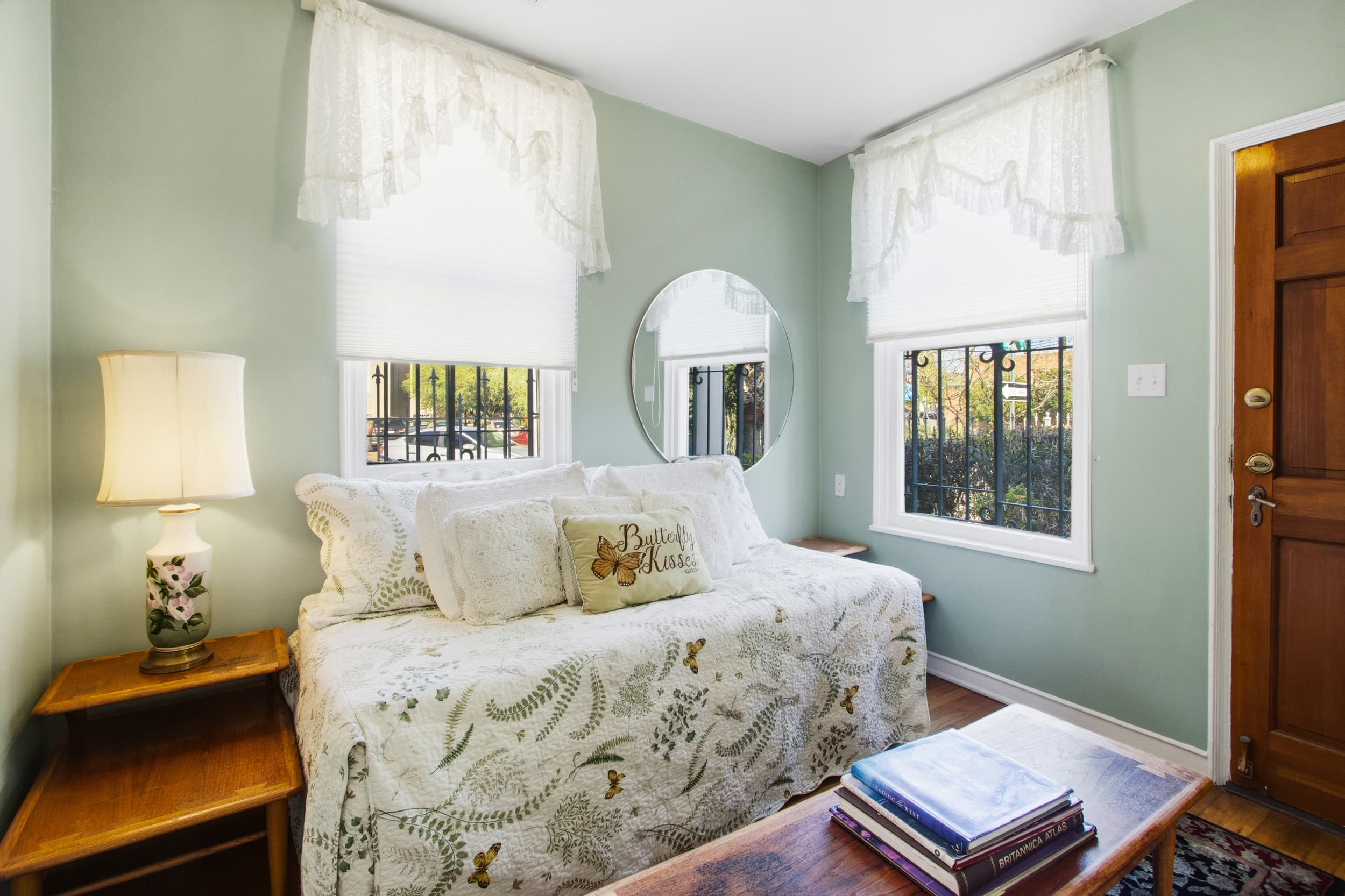 Cozy bedroom with a daybed, lamp, and large windows, adorned with light curtains and a round mirror.