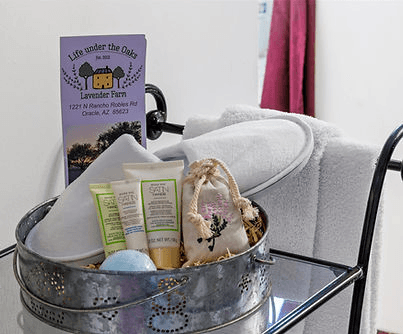 A decorative basket containing toiletries, a lavender sachet, a bath bomb, and a towel, alongside a brochure for "Life under the Oaks Lavender Farm."