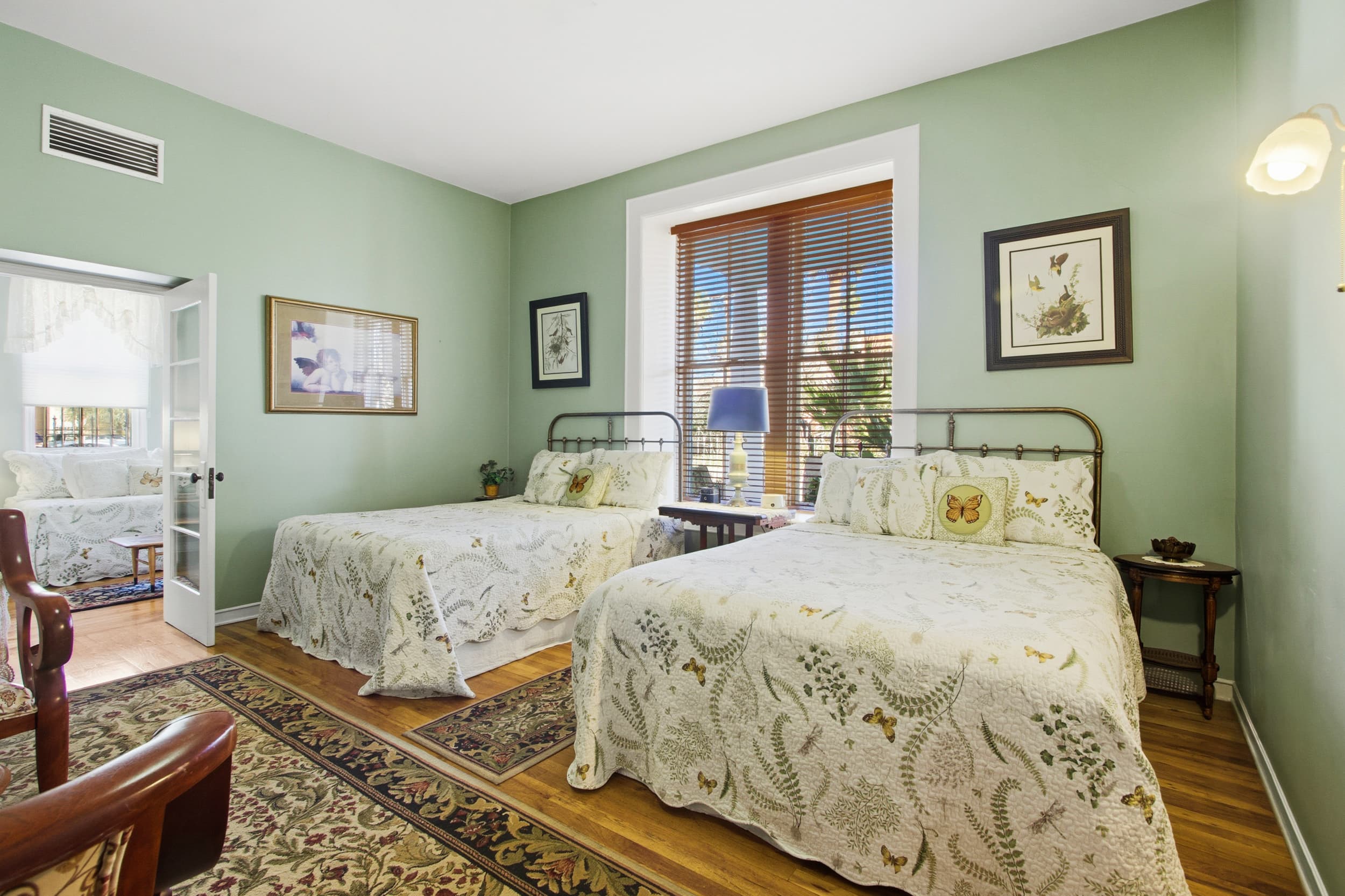 A cozy room featuring two beds with floral linens, framed artwork on green walls, and natural light from a window.