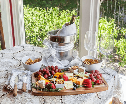 A beautifully arranged cheese platter with fruits, crackers, and a bottle of wine on a table set near a window.