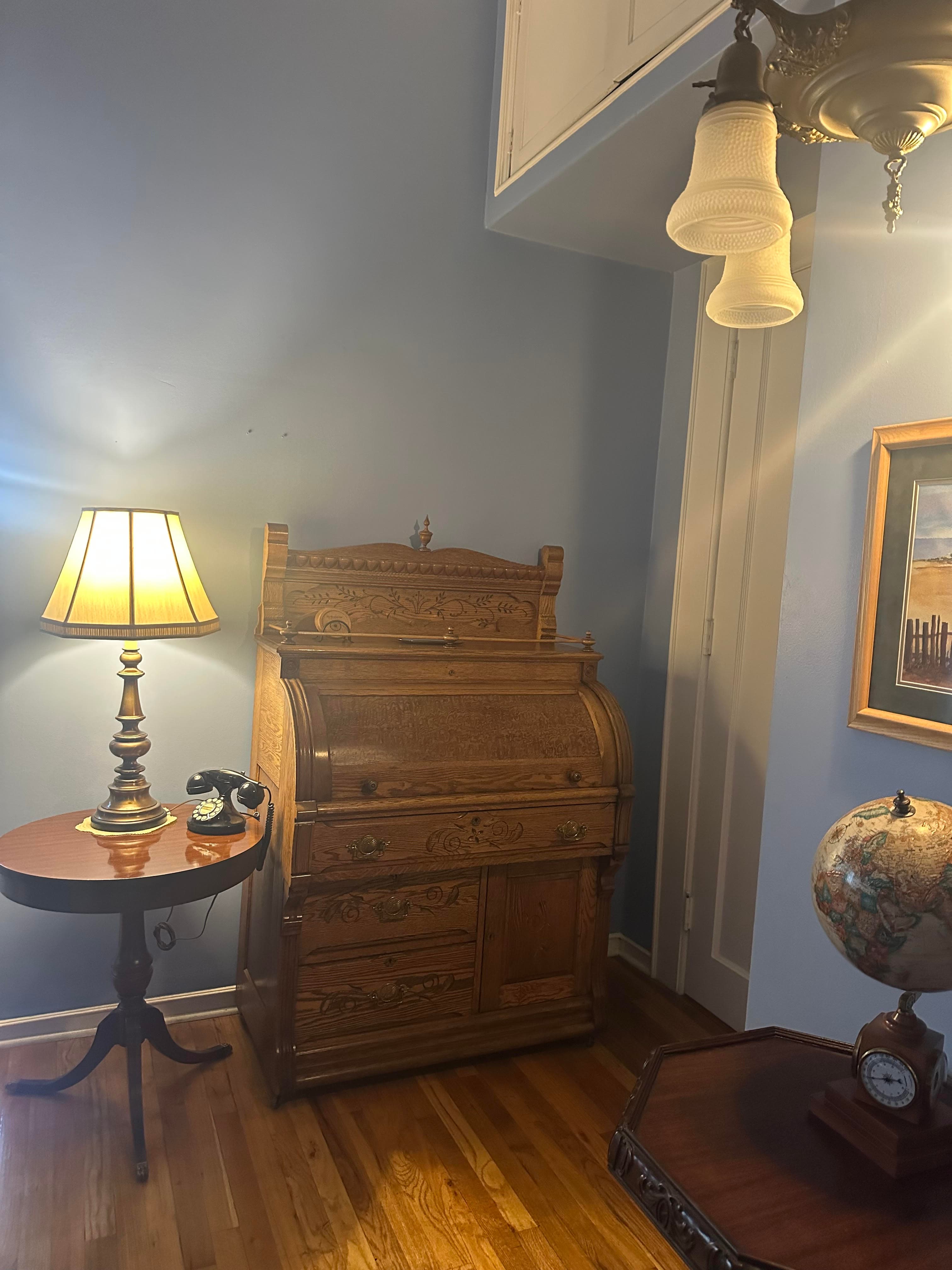 A warmly lit room featuring an ornate wooden roll-top desk, a vintage lamp, and a globe on a side table.