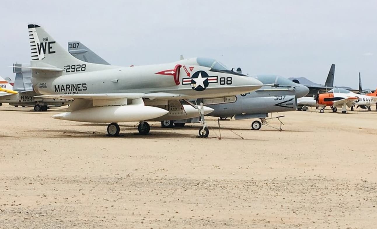 A military jet painted in gray and white, labeled "MARINES," is parked among other aircraft on a sandy surface.