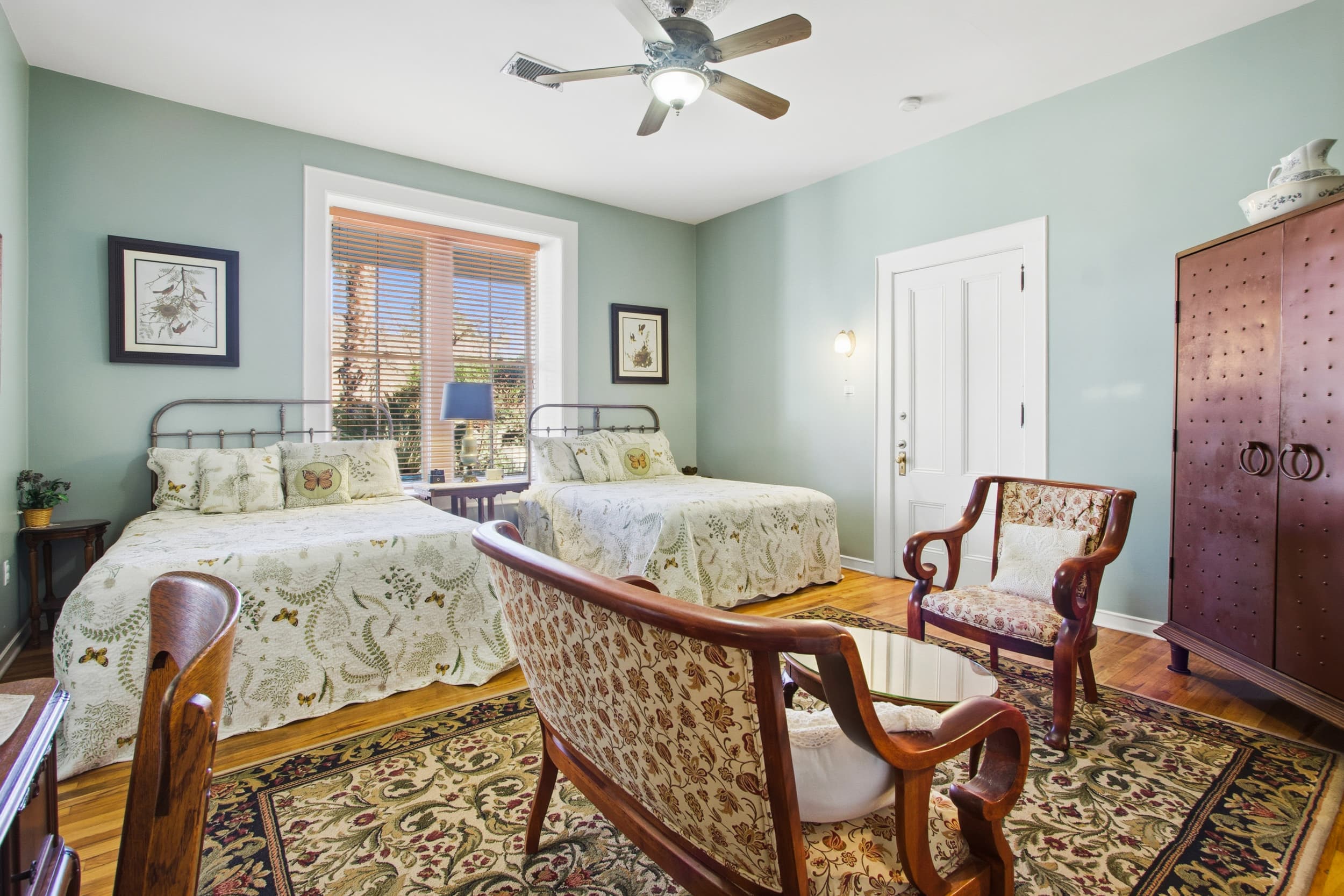 A cozy guest room featuring two twin beds, ornate seating, and natural light from a window with wooden blinds.