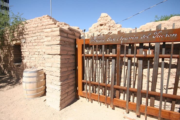 A wooden gate with a sign reading "Presidio San Agustín del Tucson" next to adobe ruins and a barrel in a sandy area. A wooden gate with a sign reading "Presidio San Agustín del Tucson" next to adobe ruins and a barrel in a sandy area.