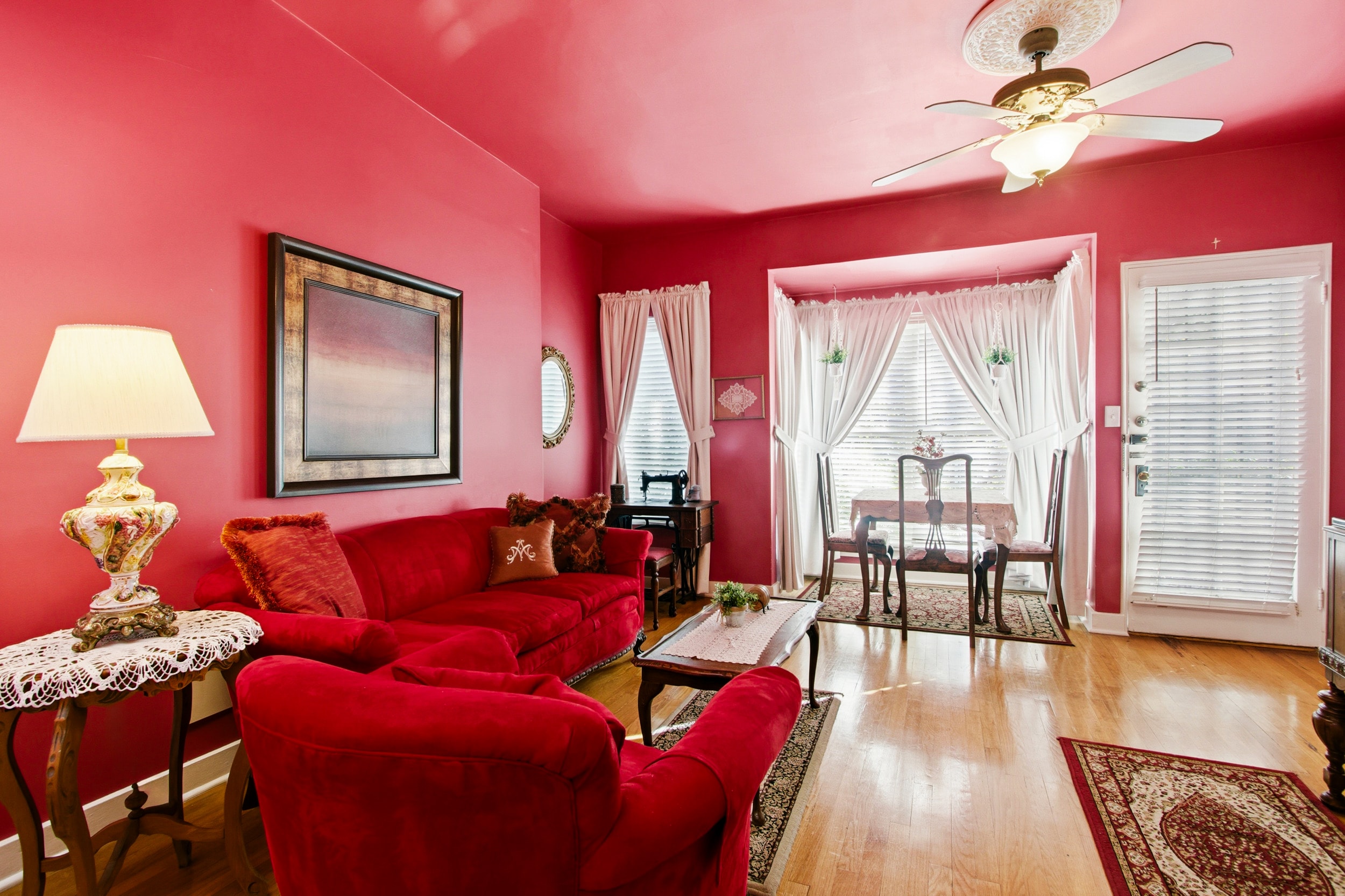 A cozy living room featuring red walls, a red couch, decorative lamps, and a dining area with large windows.